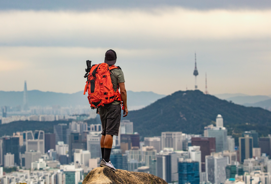 Urban backpacker standing on a mountaintop looking at a city below him.